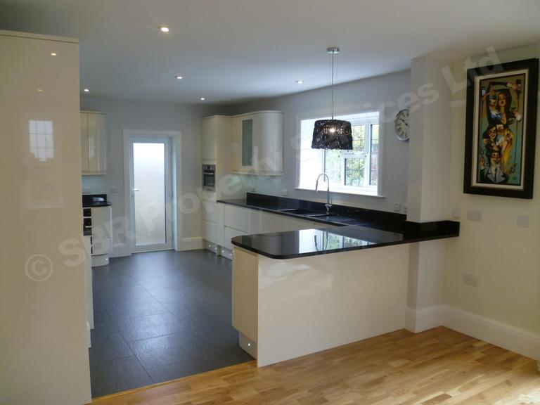 Stunning high-gloss cream kitchen in Exeter with a black granite worktop, bespoke handmade hood, hand-blown glass splashback, and range cooker by SBR Property Services Ltd.